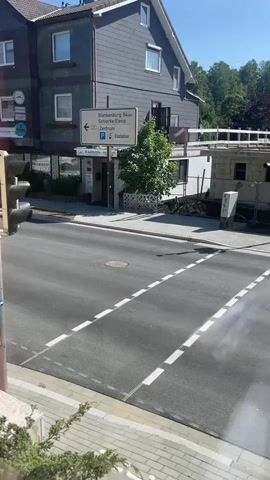 The video shows an empty street in a town on a sunny day. The camera pans from left to right, revealing buildings, a construction site, a traffic light, and street signs. There are no people or moving vehicles visible. The street is marked with white lines. The buildings are made of brick and stucco, with pitched roofs. The environment is peaceful and quiet.