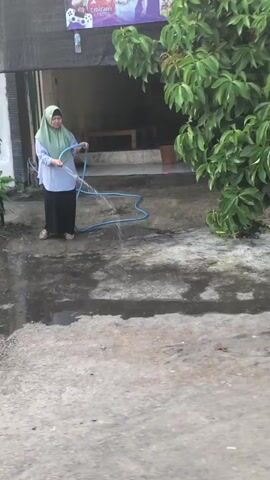 A person wearing a light blue headscarf and a light blue tunic over a dark skirt stands in a paved outdoor area, holding a blue hose from which water is spraying onto the ground. To the left, a gray trash can sits near a leafy plant. Behind the person, a building with a wooden door and windows is visible. The ground is wet in patches. The lighting suggests it is daytime, possibly overcast.
