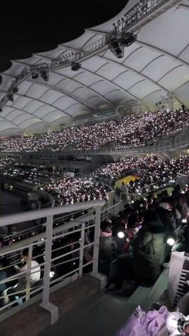 The video shows an audience at a concert venue at night. Many attendees hold illuminated devices, creating a sea of lights under the venue's arched roof. The video focuses on a section of the audience. Some individuals are seated, while others are standing. A woman films the concert with her phone. As the video progresses, the perspective shifts, and several attendees are seen waving their light sticks. They look excited and are smiling and laughing. The general atmosphere appears festive and energetic.