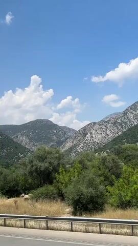 The video shows a scenic view of mountains covered in vegetation under a clear blue sky with scattered clouds. The camera appears to be moving, possibly from a vehicle, as indicated by the presence of a guardrail and a road surface in the foreground. The mountains are rugged with visible rocky outcrops and dense green foliage. The lighting suggests it is daytime, likely midday or early afternoon. The overall impression is a natural, mountainous landscape during fair weather.