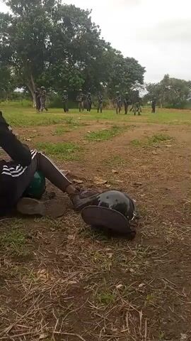 In the foreground of the video, a person is lying on the ground, partially visible from the torso down, wearing dark pants with white stripes and dark shoes. Beside them, on the ground, are two dark helmets and a dark bag. The person's right hand is visible, near the helmets. In the background, a group of approximately ten individuals, dressed in military-style uniforms and wearing green helmets, are standing in a line or scattered in a field. They appear to be holding objects, possibly rifles, and are looking in various directions. The environment is an outdoor, grassy area with some dirt patches and trees in the background. The sky is overcast.