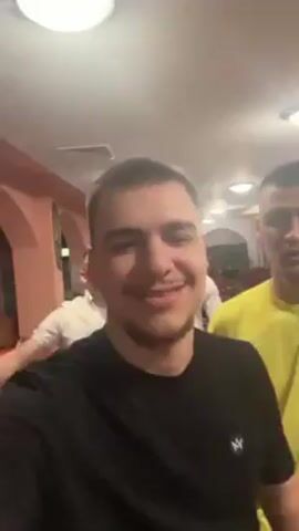The video shows a group of four young men, probably in their 20s, in what appears to be a restaurant or event venue. The man in the foreground, wearing a black t-shirt, is holding the camera in selfie mode. He is smiling and pointing at himself. Behind him are three other men. One is wearing glasses and a white button-down shirt, another is also wearing a white button-down, and the third is in a yellow shirt. They appear to be posing and having fun, with some of them raising their arms and making gestures. The venue is brightly lit with overhead lights. Tables are visible in the background, with what seem to be place settings, suggesting that it might be a banquet or party. A woman's head is visible in the background sitting at a table. There's music playing in the background.