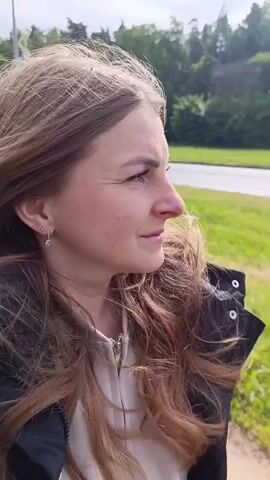 A woman with long, light brown hair smiles at the camera as her hair blows in the wind. She is wearing a beige top and a black jacket with fur lining.  The background shows a green field, a road, and trees. The weather appears overcast.