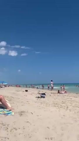 The video shows a sunny beach scene with a clear blue sky and light waves lapping on the shore. Many people are scattered across the beach and in the water, enjoying the day. The camera pans slowly across the scene, starting from a general view of the beach and then focusing on a group of people sitting under a purple beach umbrella. Beach chairs, towels, and a bag are placed around the umbrella. After focusing on the people under the umbrella, the camera pans back to show the wider view of the beach and ocean.