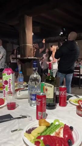 The video shows a lively party scene in what looks like a restaurant. In the foreground, a table is laden with food, including sliced vegetables and cold cuts, along with bottles of alcohol like Finlandia vodka and Jameson whiskey, cans of Coca-Cola, and a carton of Yoocy sour cherry juice. In the background, several women are dancing on a slightly elevated platform, with one woman in a black dress leading the dance and a cameraman recording the event. The venue appears to have a rustic interior with wooden pillars and beams. People are scattered around the room, some observing and others possibly socializing. The atmosphere is festive, suggesting a special occasion or celebration.