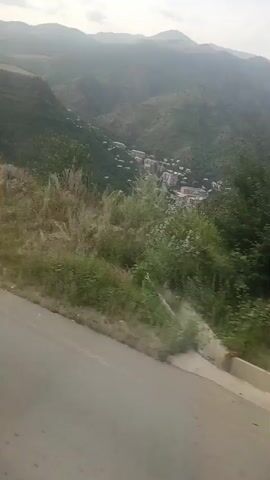 The video shows a scenic view from a moving vehicle. The camera is pointed out a window, capturing a landscape with rolling hills and a valley. There is a road in the foreground with a concrete barrier on the right side. Lush green vegetation covers the hills, and some white wildflowers are visible. In the distance, a small town or village is nestled in the valley, with buildings and houses spread out among the trees. The sky is overcast, suggesting a cloudy or hazy day. The overall impression is of a picturesque mountain or rural environment.