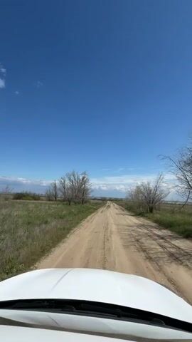 The video shows a view from the hood of a white car driving down a dirt road. The road is long and straight, leading towards the horizon where a body of water can be seen in the distance. On either side of the road, there is low green vegetation and scattered bare trees. The sky is blue with some white clouds. The car is moving forward, kicking up dust from the road.