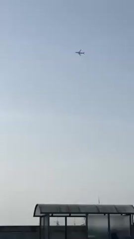 The video shows an airplane in the sky. The sky is light blue and a little hazy. At the bottom of the frame, there is a bus shelter and a tall building. The airplane is high in the sky, but it is still visible. The video is shot from a low angle.