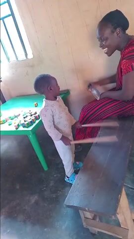 In a simple, well-lit room, an adult woman and a young child are engaged in a playful musical activity. The woman, wearing a red and black striped dress and a black head covering, is seated on a wooden bench, playfully tapping a small plastic water bottle with her hand. The child, dressed in a light-colored long-sleeved top with cloud patterns, light-colored pants, and blue clogs, stands facing the woman. The child holds two drumsticks and enthusiastically taps them on the wooden bench, mimicking drumming.

Both the woman and the child are smiling broadly and looking at each other, indicating a joyful and interactive moment. The child even lets out a laugh at one point. Natural light streams in from a window on the left side of the frame, illuminating the room. On a green plastic table next to the child, numerous colorful letter blocks are scattered, displaying letters such as 'A', 'B', 'D', 'G', 'I', 'L', 'M', 'O', 'P', 'R', 'S', 'T', 'U', 'Y'. The room features light-colored walls and a concrete floor. The overall scene suggests a heartwarming and educational playtime activity in Barakin Columbai, Nigeria.