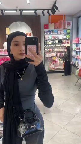 A young woman is standing in a store, taking a selfie in a mirror. She is wearing a black hijab, a long-sleeved black shirt, a dark gray vest, and dark gray pants. She has a small black purse with a strap slung across her body. The woman is holding a cell phone in her right hand. The phone has a pink case with a grid pattern. The store has shelves stocked with various items, including cosmetics, trinkets, and gifts. The lighting in the store is bright. Other shoppers are visible in the background, one wearing a hijab and a black leather jacket.
