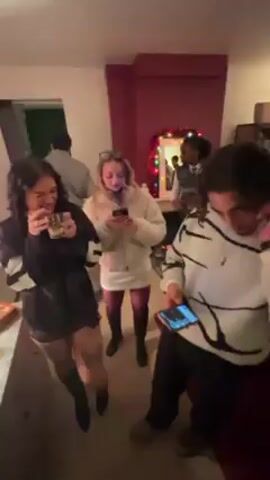 The video shows an interior scene of a house party with festive decorations. The first shot features three individuals: a woman in a black outfit holding a drink, a woman in a white dress using a phone, and a man in a patterned sweater also looking at his phone. They are standing in a room with a red accent wall adorned with a mirror and Christmas lights. The camera then pans to reveal another room with a white couch, string lights on the wall, and a group of men playing pool. The individuals are socializing and casually interacting within the house party setting.