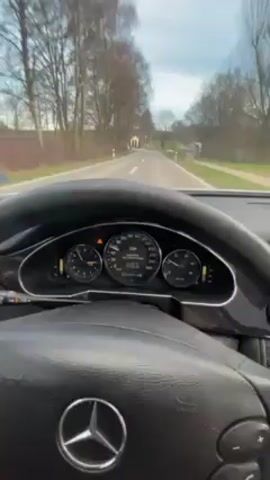 The camera is positioned inside a Mercedes-Benz, facing the windshield and the road ahead. The car is driving on a paved road through a rural area with trees and houses on either side. The dashboard and steering wheel of the car are visible. The speedometer shows the car is moving at approximately 50 kilometers per hour. The sky is overcast. There is music playing in the background. The camera is mostly static, with occasional slight movements due to the car's motion.