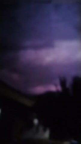 The video begins with a dark, blurry frame. The scene appears to be outdoors at night. A faint light source is visible on the left side of the frame, possibly a window or a lamp. In the center of the frame, a dark, amorphous shape suggests the presence of trees or foliage. The overall ambiance is dim and indistinct.