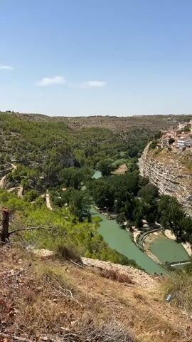 A video shows a scenic landscape of a lush green valley with a river running through it. The river has a distinctly turquoise hue and is lined with abundant trees. On the right side of the valley, a town is built into a cliff face, with buildings clustered together and a prominent church spire visible. The terrain is hilly and dry, with patches of vegetation and sparse trees on the slopes leading up to the valley. The sky is clear and blue, indicating a sunny day. There are no people or discernible actions in the video, making it a static landscape shot.