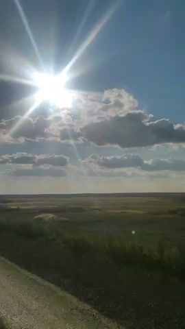 THE VIDEO SHOWS A DIRT ROAD ON THE LEFT AND A FIELD WITH GRASS AND SOME TREES ON THE RIGHT. THE SUN IS SHINING BRIGHTLY IN THE SKY, AND THERE ARE SOME CLOUDS. THERE IS A ROUND SIGN ON A POLE IN THE FIELD. THE VIDEO IS TAKEN FROM A MOVING VEHICLE.