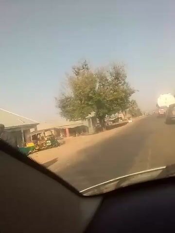 The video shows a view from inside a car driving along a road in a rural area on a sunny day. A tree stands on the side of the road next to a small building. There are other structures and a water tower visible in the distance. Other vehicles, including a tanker truck, are also on the road. The area appears dusty or hazy. The camera angle is slightly tilted.