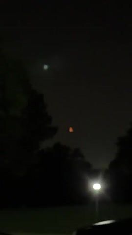 The video shows a night sky with a crescent moon appearing orange. There are trees silhouetted against the sky, and a light fixture is visible in the lower right corner. The moon is the primary focus, and its color changes slightly throughout the short clip.