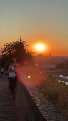 The video opens with a group of people standing on a brick walkway, looking out at a sunset over a city. The sun is a bright, orange orb in the sky, casting a warm glow over the buildings below. The people are silhouetted against the sunset, their faces obscured by the light. The camera pans slightly, showing more of the city and the river that runs through it. The sky is a vibrant orange, fading to a pale yellow at the top of the frame. A few trees are visible in the foreground, their leaves dark against the bright sky. The people on the walkway appear to be enjoying the view, with one person holding up a phone as if to take a picture. The scene is peaceful and serene, capturing a beautiful moment at the end of the day.