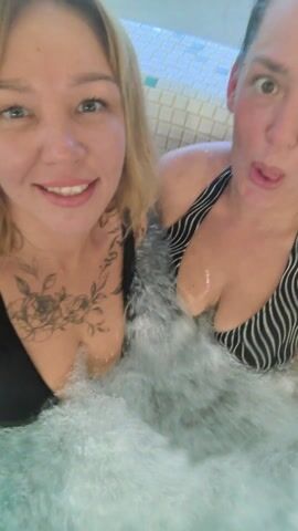 Two women are in a hot tub. The woman on the left has blonde hair, fair skin, and a black bikini top. She has a floral tattoo on her chest. The woman is smiling and looking at the camera. The woman on the right has brown hair and is wearing a black and white striped bikini top. She is making faces at the camera. The hot tub is filled with bubbling water. The sides of the hot tub are tiled with white and blue mosaic tiles.