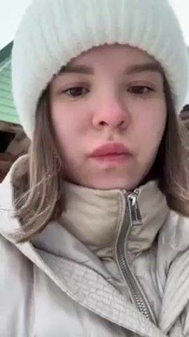 A young woman with fair skin, brown eyes, and light brown hair is looking directly at the camera. She is wearing a white knit hat and a light-colored puffer jacket zipped up to her neck. She puckers her lips slightly. In the background, a building with green trim can be partially seen. The scene appears to be outdoors, with possible snow or light-colored debris on the ground. The shot is a close-up, focusing on the woman's face.