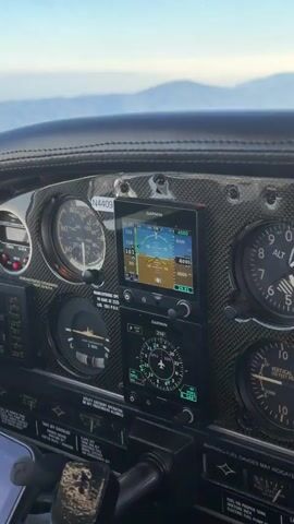The video shows a close-up view of an airplane's cockpit instrument panel. The camera pans across various gauges and screens, providing a detailed look at the flight instruments. The primary focus is on the digital displays, which show flight information such as altitude, airspeed, heading, and navigation data. Traditional analog gauges, including an altimeter and a vertical speed indicator, are also visible. The dashboard appears to be made of a dark, textured material, possibly leather or carbon fiber. The lighting suggests it is daytime, with natural light illuminating the cockpit. The background is out of focus but appears to be a landscape with hills or mountains and a clear sky. The overall impression is one of an operational aircraft cockpit, with the instruments actively displaying data.
