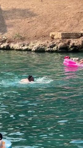 A man is swimming in clear turquoise water. He is in the foreground and appears to be doing the front crawl. In the background, a pink inflatable mattress with two people on it is floating. One person is wearing a blue swimsuit and a white cap, the other is partially obscured. Further to the right, another person in a yellow floatation device is in the water, and a man in a dark t-shirt is standing on the edge of the water. The water is calm with small ripples. The background shows a sandy bank with rocks and vegetation. The lighting suggests it is daytime.