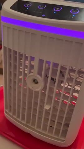 The video shows a close-up of a white air cooler or fan with blue and purple lights. The device is placed on a red surface. The top of the device features several buttons with illuminated icons, and some visible text that appears to be a timer setting: "2H 4H 6H". The fan blades are visible through the white grille, and the lighting from the device itself casts a glow on its surroundings. The scene is indoors, likely a room, and the overall impression is of a functional, modern appliance. The lighting suggests it might be evening or nighttime. There are no people or animals visible in the frame. The context of Varese, Italy, does not provide specific visual cues within the video itself.