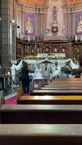 A wedding ceremony is taking place inside a church. A bride in a white wedding dress and groom in a light-colored suit stand at the altar, facing away from the camera. A officiant in white robes stands to the right of the couple. Two figures in dark clothing, likely part of the wedding party, stand to the left of the couple. The altar is decorated with white fabric drapery and floral arrangements. Pews filled with wedding guests are visible in the foreground, with rows of wooden pews facing the altar. The church interior features ornate architecture, including painted murals on the ceiling and walls depicting religious figures, and elaborate chandeliers. The overall atmosphere suggests a formal religious ceremony.