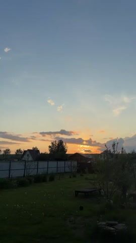 This video captures a dusky outdoor scene, likely a backyard, as the sun begins to set. The sky is a gradient of blues and oranges, with scattered clouds catching the last rays of sunlight. Silhouetted against the sky are trees, a fence, and parts of houses, suggesting a rural or suburban environment. The foreground is dark, with the details of the ground and any objects on it obscured by shadow. The camera angle is tilted upwards, focusing on the sky and the distant landscape. The video appears to be a time-lapse or slow-motion recording, as the clouds and light change subtly over a few seconds. No people are visible in the video.