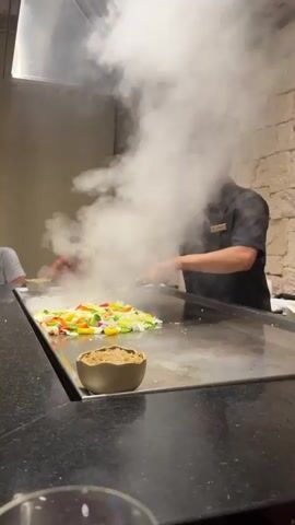 A male chef in a black uniform and chef hat is cooking vegetables on a large flat-top grill. There is a lot of steam rising from the grill. In the foreground, there is a small brown bowl with what appears to be rice or some kind of topping. Behind the chef, two people are seated at the grill, also watching the cooking. The background appears to be a restaurant setting with stone-like walls. The chef is actively cooking the vegetables with spatulas, flipping them and stirring them.