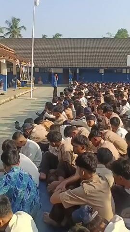 A vertical video shows a large group of students, all appearing to be male and dressed in school uniforms, sitting on the ground in rows. They are outdoors in a schoolyard, with buildings and a basketball hoop visible in the background. The students are facing a man standing in front of them, who is speaking. The students are mostly looking down, some are holding papers, and some are interacting with each other. The sun is shining, indicating it is daytime.