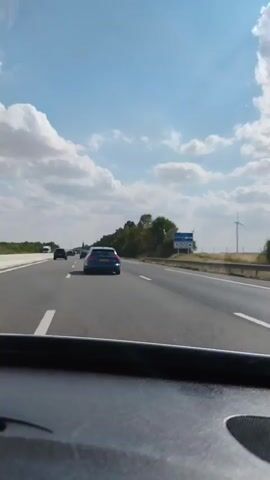 The video captures a view from inside a vehicle traveling on a highway under a partly cloudy blue sky. The road has multiple lanes, and several other cars are visible, including a blue SUV in the foreground. Trees and vegetation line the side of the highway. In the distance, there is a wind turbine and a road sign. The camera perspective suggests the viewer is a passenger.