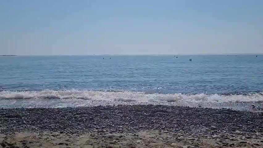 A wide shot shows a calm, blue ocean with gentle waves rolling onto a pebble beach. The sky is clear and bright blue. There are a few buoys or markers visible in the distance on the water. The camera appears to be static, facing the ocean.