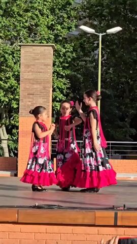 Three young girls in matching flamenco dresses are dancing on a stage. The dresses are black with a floral pattern of pink, white, and green, and have ruffled red trim. The girls have their hair pulled back in buns. They are moving their arms and hands in coordinated gestures. The stage is made of wood and is set outdoors, with trees and a brick pillar in the background. A lamppost is visible above the trees. The lighting suggests it is daytime.