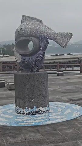 A video of a sculpture depicting two fish, one on top of the other, with a circular opening in the middle. The sculpture is made of mosaic tiles and stands on a cylindrical base. In the background, a person is playing a flute. The scene appears to be outdoors, with a body of water and a breakwater in the distance. The weather is overcast.
