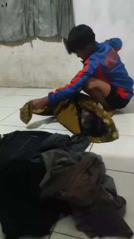 A young boy crouches on a tiled floor, sorting through a pile of clothes. He is wearing a blue and red long-sleeved shirt and black shorts. In front of him is a large pile of clothes, including black, grey, brown, and a gold patterned garment that he is holding. In the background, there is a wall with a grey curtain.