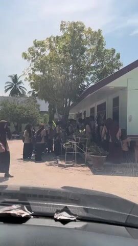 The video shows a group of students, many wearing hijabs, walking on a dirt road near a green school building. Some students are carrying backpacks. A tree provides shade, and a few plants are in pots near the building. The footage is filmed from inside a car, as the dashboard is visible at the bottom of the frame. The day appears sunny.
