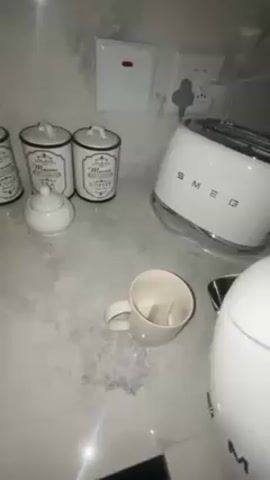 The video shows a person pouring water from a white electric kettle into a white mug. Two tea bags are already in the mug. The mug is on a countertop. Behind the mug, there is a white Smeg toaster, three white ceramic containers with black lettering that say "Sugar", "Paris Maison", and "Coffee", and two electrical outlets. One of the outlets has a plug in it. There is also a light switch. The countertop is a light grey color.