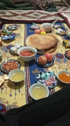 The video shows a spread of food laid out on a patterned cloth on what appears to be the floor. The cloth has a yellow background with blue and gold patterns.  There are several small bowls and plates containing various food items. These include a flatbread, a bowl of red sauce, fruit (apples and peaches), candies, and small bowls of light and dark liquids. A clear pitcher with an orange liquid inside is also visible. A teapot and some packaged snacks are near the pitcher.  A person is visible in the lower right corner, wearing dark pants.  The camera moves slightly and there is a brief, blurred glimpse of a hand reaching towards the food.  There is someone speaking in the background, but the audio is indistinct. A red and brown patterned blanket is visible on a couch in the background.