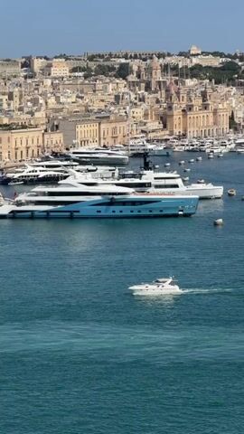 The video shows a harbor with several large yachts docked in the background. A smaller white motorboat is moving through the foreground of the turquoise water. The city skyline, characterized by beige and cream buildings with a few dome-topped structures, is visible in the background under a clear blue sky. The overall scene depicts a sunny day at a bustling port with luxury vessels.