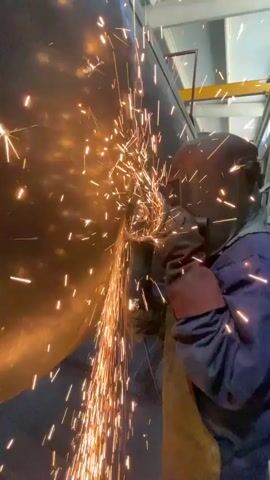 A person wearing a blue jumpsuit, a dark face shield, and red gloves is grinding a large metal surface with an angle grinder. Sparks are flying off the metal in a shower of orange light. The background appears to be an industrial setting, with exposed beams and lighting fixtures visible overhead. The metal surface being worked on is dark and appears to be curved. The grinder is being held with both hands.