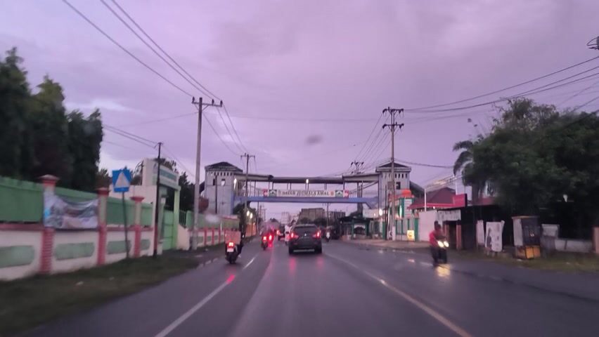 The image displays an inverted view of a street scene, likely captured during dawn, dusk, or an overcast day, given the soft, ambient lighting and the wet road surface. The perspective makes the sky appear as the bottom half of the frame and the road as the top.

A prominent, ornate archway spans across what would be the road (at the top of the inverted image). This archway features two tower-like structures on either side and is adorned with banners. The most legible text on the archway reads "DAYAH JEUMALA AMAL" and "AKREDITASI 'A'". A red banner also says "SELAMAT DATANG DI" (Welcome to). These texts suggest an Indonesian setting, likely indicating an entrance to a religious educational institution or a specific area named after it.

The road itself is made of asphalt, appearing wet and reflective. It has visible white pedestrian crossing lines and a yellow median line. Several vehicles are present, including cars with illuminated taillights and motorcycles. One motorcycle rider is visible on the left side of the inverted road.

Along the sides of the road, various buildings are seen, resembling shops or small commercial establishments. On the right side, a green and white fence runs alongside the road. Utility poles with a dense network of overhead wires are conspicuous, particularly in the lower (sky) portion of the inverted image. Vegetation, including palm trees and other greenery, is scattered throughout the scene. Other signs, such as a white sign with "BSI" (likely Bank Syariah Indonesia) on the left and a blue directional arrow sign on the right, are also visible. The sky, appearing at the bottom, is a muted, pale purple-gray, reinforcing the low-light conditions.