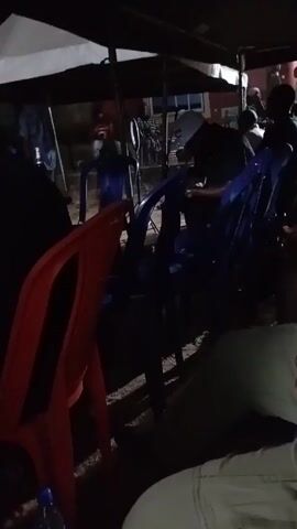 The video shows a gathering of people under a tent at night. Several plastic chairs, colored red and blue, are arranged around the tent. Individuals are seated, though their features are largely obscured by the darkness. One person in a blue outfit is walking around. Music is playing in the background, suggesting a social event or celebration. The setting appears to be outdoors, with a building visible in the background.