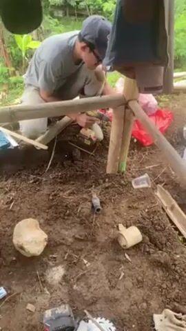 A young man in a grey shirt, tan pants, and a dark baseball cap is kneeling and using a power drill to work on a bamboo structure set into the soil.  The structure appears to be a frame or simple shelter, with bamboo poles forming a lattice.  The soil around the structure is disturbed and littered with debris, including a rock, plastic cups, a metal can, and sections of bamboo.  A red plastic bag sits behind the man, partially obscured by the bamboo frame.  A jacket or blanket hangs from the structure above him. The background shows a lush green hillside with trees and foliage.  The scene is shot from a slightly elevated angle, looking down into the work area.
