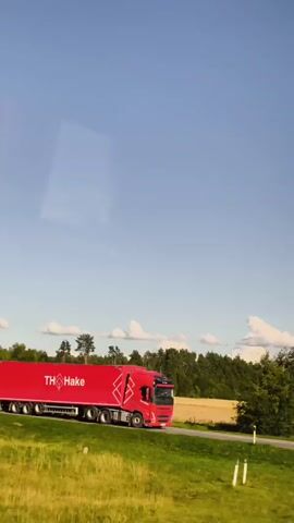 A large red truck with the logo TH Hake on its side travels down a paved road. Beside the truck, a smaller black car is driving in the same direction. The road is bordered by a grassy field and a line of trees in the background. The sky is clear and blue with a few scattered clouds. The video is shot from the perspective of someone inside a vehicle moving alongside the truck and car.
