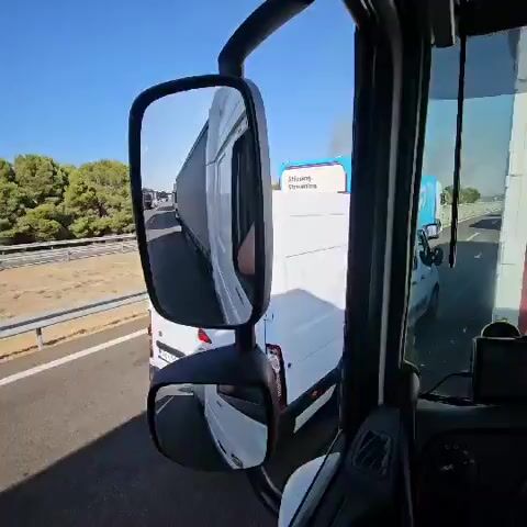 The video shows a view from inside a truck cab, looking out the driver's side mirror. The mirror reflects a line of traffic, primarily consisting of large trucks and some cars, moving along a highway. The time stamp indicates the video is approximately five seconds long. The camera appears to be mounted inside the cab, possibly on the dashboard or windshield, as it also captures parts of the truck's interior, including the side of the driver's seat and a glimpse of a person's leg. The environment appears to be a sunny day, with a clear blue sky and vegetation visible alongside the highway. The traffic is either stopped or moving very slowly, suggesting a potential traffic jam or congestion. One of the reflected trucks in the mirror has a blue and white advertisement on its side that is partially visible. The overall scene depicts a common highway travel situation with traffic delays.