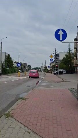 A pink car is driving down a residential street under a cloudy sky. The street is paved with asphalt and has a sidewalk made of red bricks. There are several traffic signs visible, including a blue sign with a white arrow pointing straight ahead and a yellow diamond-shaped sign with a black pedestrian symbol. A white car is parked on the right side of the street, and there are buildings on both sides of the road. The video appears to have been recorded from a moving vehicle.