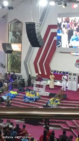 The video shows the interior of a church or large meeting hall.  The focus is on the stage area, which is decorated with flowers, and has a red-carpeted tiered platform.  Three people are on stage: two are standing behind a white podium, and one is standing to the side. All are dressed in traditional African clothing. Behind them is a large screen displaying a crowd of people and a red and white striped architectural design. Speakers are suspended from the ceiling. The hall is full of people seated and watching. The video is taken from a lower angle within the crowd.