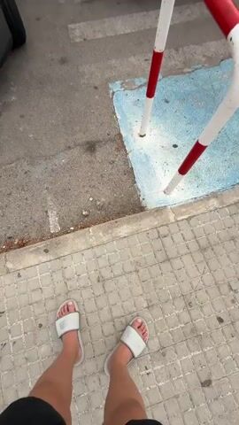 The video begins with a close-up shot of feet wearing white slides on a tiled sidewalk. The camera then pans up and to the right, revealing a gray car parked on asphalt next to a blue van. A red and white bollard is visible in the foreground. The view expands to show a large white building in the background, possibly a hotel, with palm trees and other greenery. Several cars are parked in a row in front of the building, including a red SUV. The sky is overcast with some sunlight breaking through. The license plate on the blue van reads "4039 JDG". The video captures an outdoor scene in what appears to be a parking lot or street in front of a building.