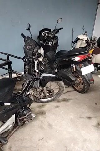 The video shows an outdoor scene with several motorcycles parked. In the foreground, a black motorcycle is visible, with its front wheel turned to the left. Behind it, a white and maroon motorcycle is parked. Further back, a black scooter is parked, and to its right, a white vehicle, possibly an ambulance, is parked. The ground appears to be concrete. The lighting suggests it is daytime.